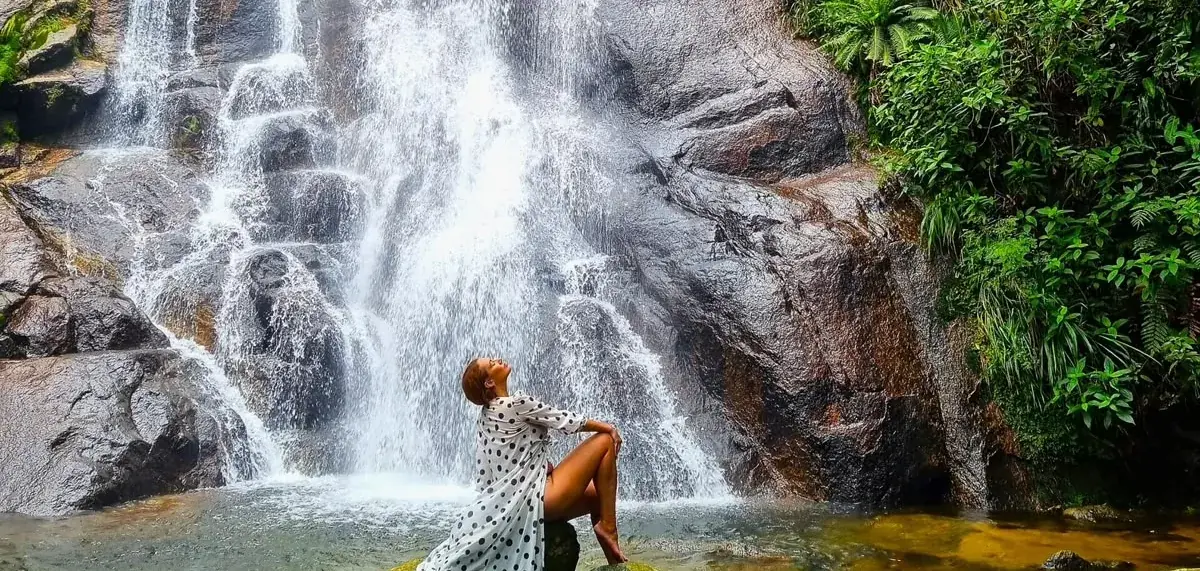 Mulher sentada numa pedra diante de uma cachoeira alta em meio à mata em Ilhabela
