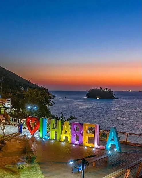 Letreiro Amo Ilhabela iluminado no Mirante do Piúva ao pôr do sol, com a Ilha das Cabras ao fundo