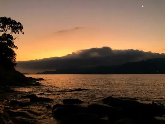 Pôr do sol visto das pedras da Praia do Oscar, com a Serra do Mar ao fundo
